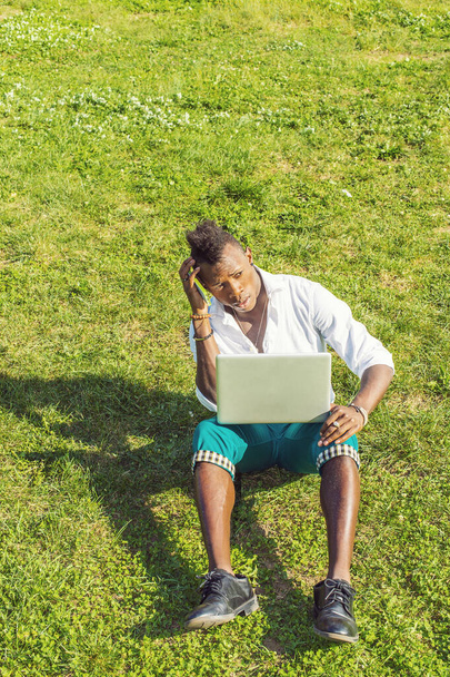 Vestido com uma camisa branca, calças verdes, sapatos de couro preto, segurando um computador portátil e um telefone celular, arranhando a cabeça, um jovem negro com cabelo moicano está sentado em uma grama verde, pensando - Foto, Imagem