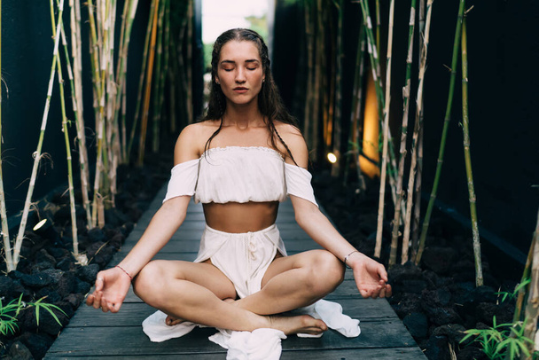 Content female in white top sitting with crossed legs on floor in Padmasana position while practicing meditation in garden during vacation - Photo, Image