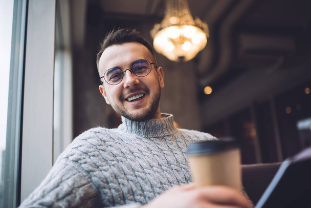 Low angle of smiling bearded male in casual wear and eyeglasses holding cup of takeaway coffee and looking at camera while chilling with book - Foto, immagini