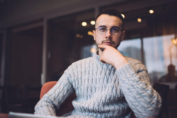 Serious bearded man in casual wear and eyeglasses sitting in comfortable armchair and looking at camera while working on laptop - Foto, afbeelding