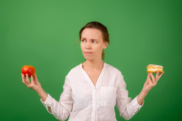 An attractive middle-aged woman in a white shirt holds an apple and a cake in each hand on a green background. The agony of choice. The concept of choosing the right healthy food. - Foto, immagini