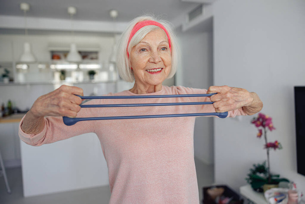 Cheerful woman pensioner exercising with elastic band at home - Foto, Imagem