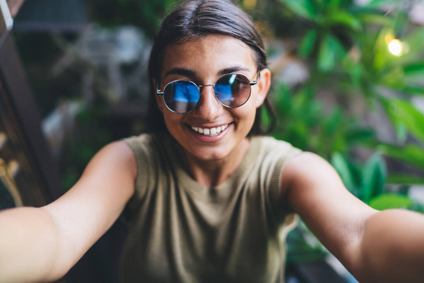 High angle of delighted ethnic female with toothy smile wearing sunglasses and taking selfie while looking at camera in tropical resort with exotic plants on blurred background - Foto, Bild
