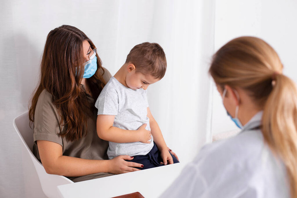 Young brunette woman with little boy having consultation at pediatrician office. Child has abdominal pain. Doctor, child and mother wearing facemasks during coronavirus and flu outbreak. Covid 19. - Photo, Image