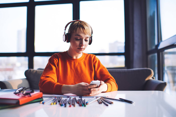 Young female designer with headphones browsing mobile phone while working at creative project sitting at table with papers colored pencils and books - Photo, image