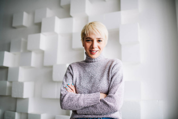 Young female in trendy clothes with short blond hair and folded hands looking at camera standing on blurred background of white wall with cube decorations - Fotografie, Obrázek