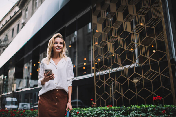 Low angle of young happy blond female in smart casual outfit messaging using mobile phone standing near modern building on street - Fotoğraf, Görsel
