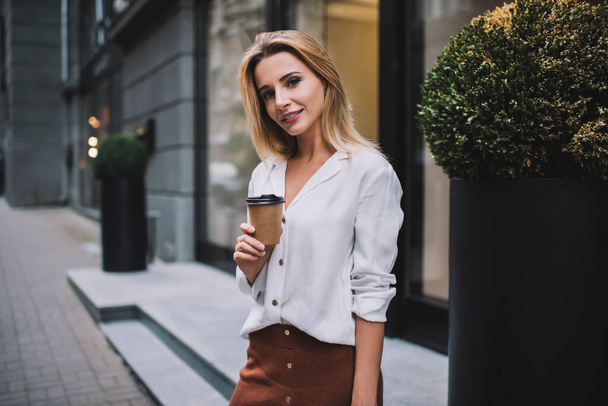 Young female in white blouse standing on pavement near entrance at shop with coffee to go and looking at camera - Foto, Imagem