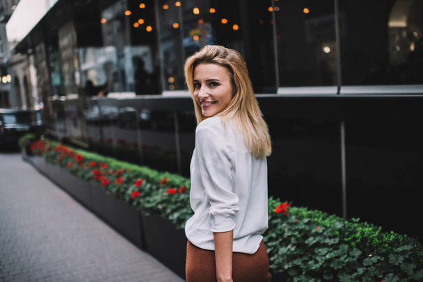 Back view of smiling blond female in white blouse walking along modern building on street and looking over shoulder in evening - Valokuva, kuva