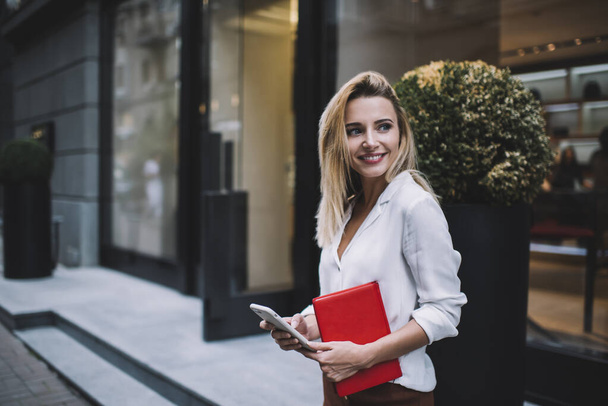 Happy young female entrepreneur in white formal blouse standing on street with planner and looking away while text messaging on smartphone - 写真・画像
