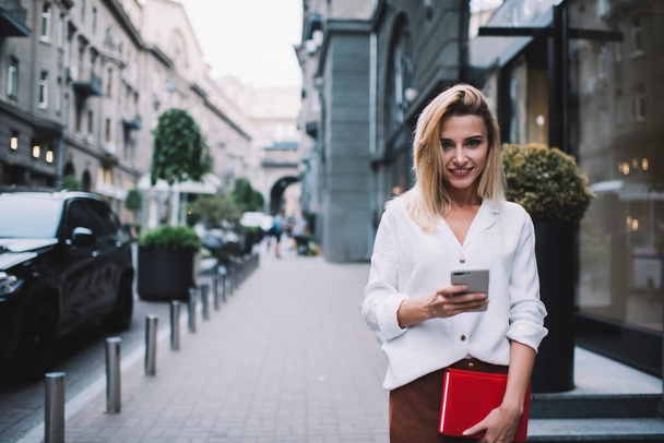 Content young female entrepreneur in formal white blouse and brown skirt standing on sidewalk near building with planner and looking at camera while messaging on smartphone - Photo, image