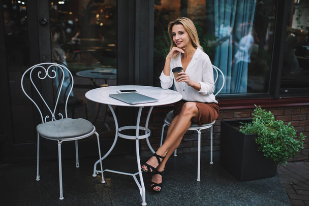 Full body happy elegant lady enjoying hot drink in paper cup while having break in remote work with gadgets in sidewalk cafe - Photo, Image