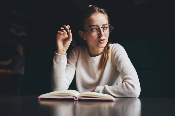 From below of concentrated female freelancer in casual clothes with eyeglasses looking away sitting at table with pen and opened notebook while working over time - Фото, зображення
