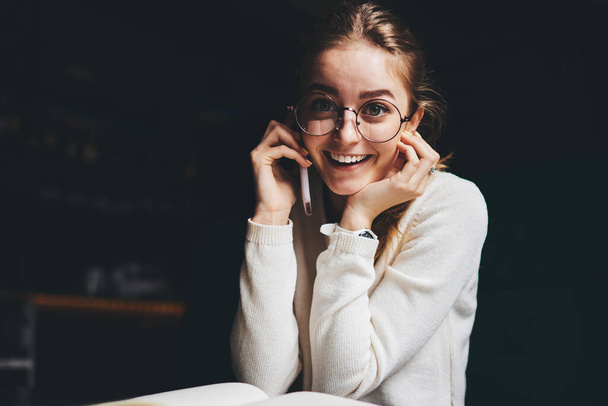 Young cheerful female in white jumper and eyeglasses discussing news on mobile phone and laughing at camera and sitting at table - Fotografie, Obrázek