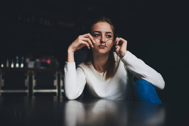 Young pensive female in white jumper jeans and round eyeglasses sitting at table and thoughtfully looking away on blurred background - Photo, image