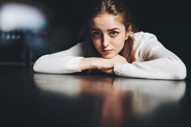 Young pensive female in white jumper sitting at table putting head on hands and looking at camera in dark room - Zdjęcie, obraz