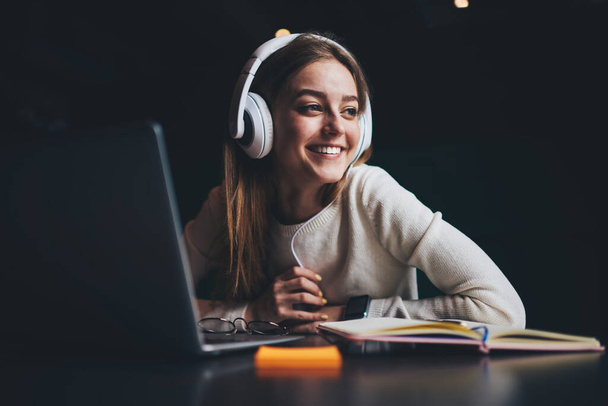 Positive female freelancer in casual outfit with toothy smile looking away and listen to music in headphones while having break in remote work on laptop in modern workspace - Foto, afbeelding