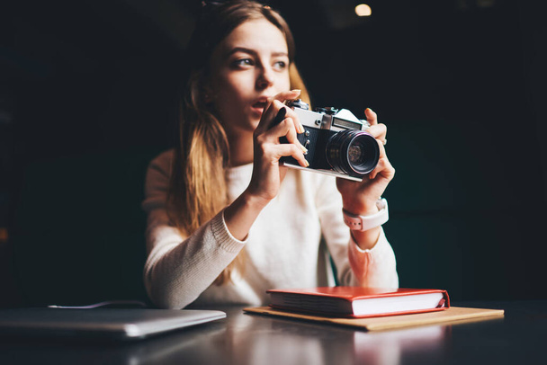 Concentrated female photographer in casual clothes looking away and taking photos on vintage photo camera with opened mouth while sitting at black table with closed laptop and notebook - Foto, afbeelding