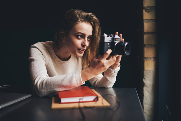 Young female in white jumper shooting with old fashioned analog camera sitting at table with book near window and looking away - Фото, зображення