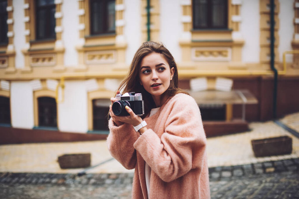 Cheerful young long haired female photographer in pink cardigan taking picture with old fashioned camera on city - Foto, Bild