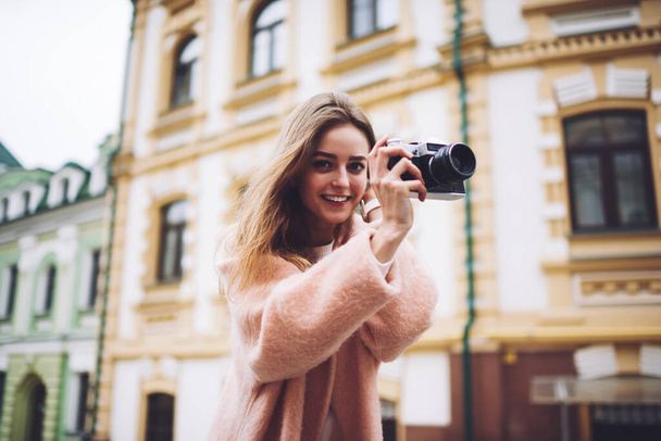 Low angle of young happy female tourist taking photo with retro camera in historical centre of city and looking at camera - Фото, изображение