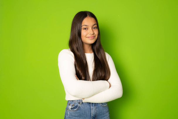 Portrait of a smiling young girl with long brunette hair in winter clothing standing with arms folded isolated over green background - Фото, изображение
