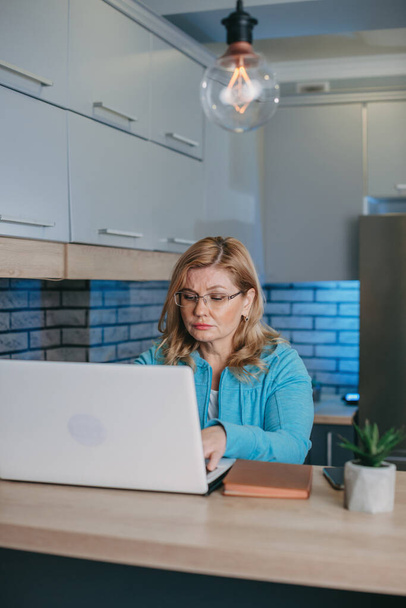 Foto de una mujer de negocios de mediana edad enfocada en gafas sentadas frente a la computadora portátil y trabajando. Estilo de vida cuarentena. Mujer de pelo rubio. - Foto, imagen