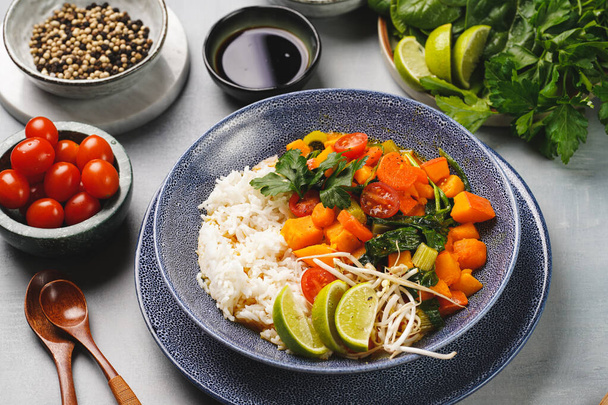 Een blauwe schaal met rode Thaise groente curry en basmati rijst, verse limoen, koriander, spinazie, chilipepers en kerstomaten - Foto, afbeelding