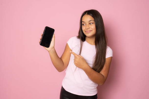 Glückliche schöne junge Mädchen mit langen Haaren hält leere Bildschirm-Handy und zeigt mit dem Finger über rosa Hintergrund - Foto, Bild