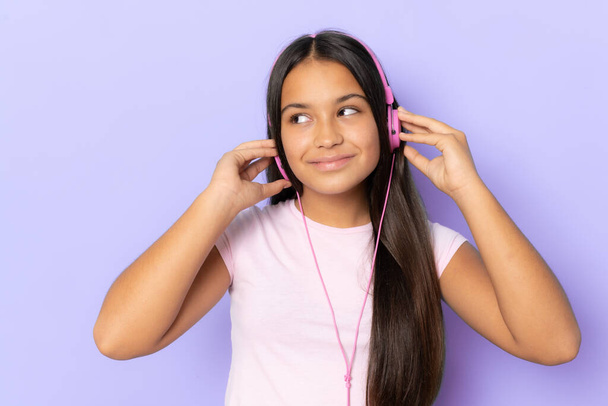 Brunette hispanic girl wearing headphones listening music over purple background - Фото, зображення