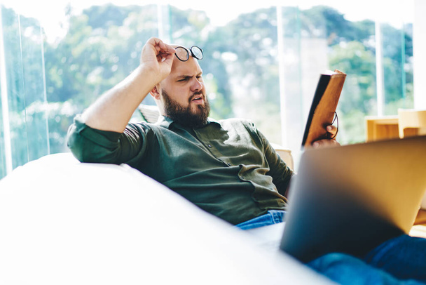 Side view of young man with beard reading book holding eyeglasses and squinting eyes for making better eyesight while resting on sofa with laptop - Photo, image