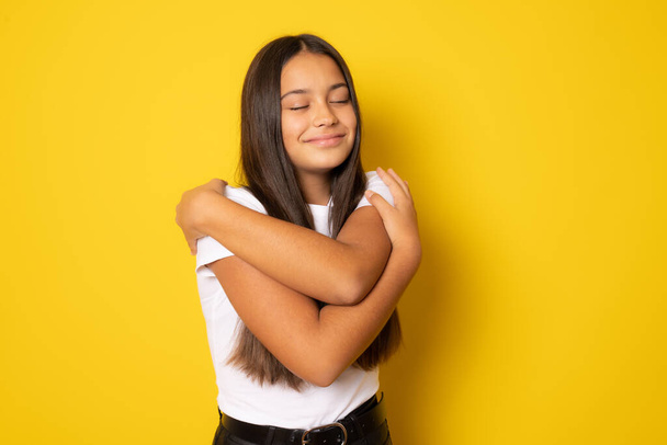 Μπρουνέτ ισπανόφωνη κοπέλα χαμογελώντας χαρούμενη αγκαλιάζοντας τον εαυτό της στέκεται απομονωμένη πάνω από κίτρινο φόντο. - Φωτογραφία, εικόνα