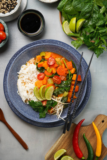 A blue bowl with red thai vegetable curry and basmati rice, fresh lime, cilantro, spinach, chilli peppers and cherry tomatoes, top view - Fotografie, Obrázek