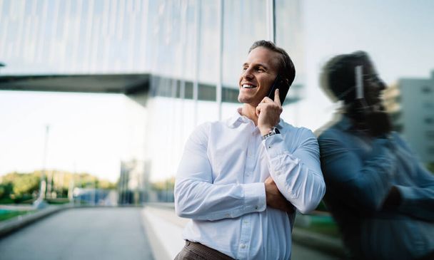 Cheerful businessman in white shirt smiling near urban building enjoying positive international conversation, happy male entrepreneur calling to friendly partner using roaming wireless connection - Foto, afbeelding