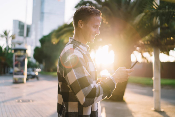 Side view of guy in casual wear standing on street smiling and using smartphone on warm summer evening in back lit - Photo, image