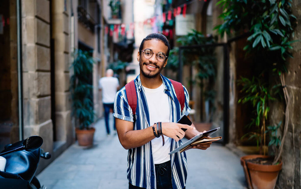 Positive young man in trendy wear with smartphone and tablet standing on street old town while checking map on tablet - Photo, image