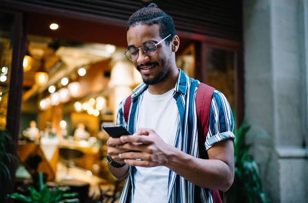 Positive dark skinned male millennial in eyewear dialling number standing on street satisfied with roaming connection, smiling african american hipster guy checking mail in social networks on street - Foto, Imagem