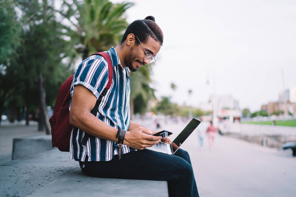 Side view of positive ethnic man in trendy clothes and eyeglasses working remotely on laptop and browsing smartphone on stone parapet on street with palms - Фото, зображення
