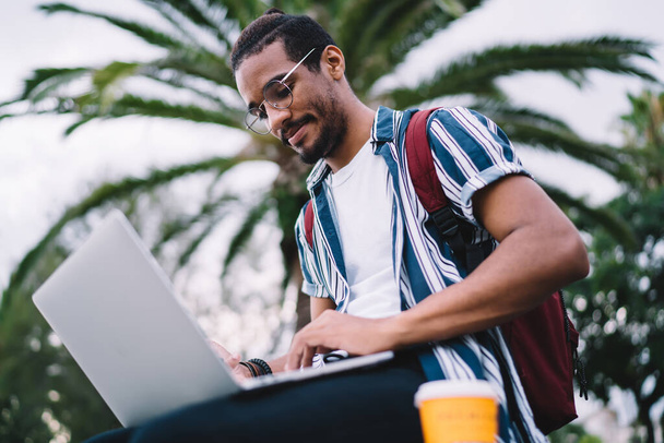 Pensive афроамериканський хіпстер хлопець в окулярах для виправлення зору ділитися публікаціями та мультимедіа в блозі, сидячи на відкритому повітрі з ноутбуком, молодий студент тисячоліття робить домашнє завдання онлайн
 - Фото, зображення