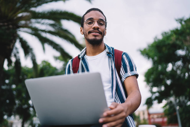 Portrait of skilled african american male freelancer in eyewear for vision correction using laptop computer for share multimedia and blogging,positive young dark skinned male student learning outdoors - 写真・画像
