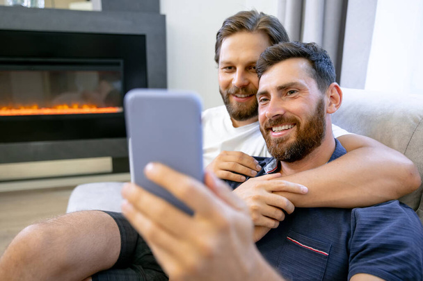 Cute male gay couple spend time at home. They lie on the couch and use a smartphone, take a selfie. The partner's hand is around his lover. The room has a modern interior. - Foto, imagen