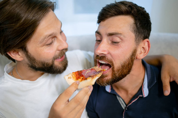 Gay marriage. The guys are in the living room, sitting on the couch with pizza. They give each other a slice of pizza. Both young and European. - Foto, Imagem