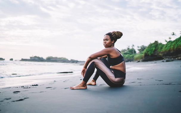 Young female athlete thinking about body positive while resting at black sandy beach during time for morning cardio workout, African American barefoot woman in tracksuit keeping healthy lifestyle - Fotografie, Obrázek