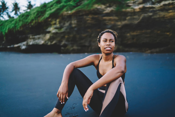 Slim woman 30s resting at black sand of seashore thinking about cardio training for have muscular body, African American female runner in sportive clothing resting during morning workout at coastline - Foto, Imagen