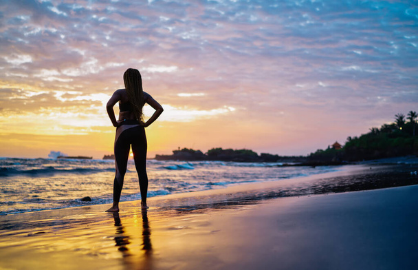 Back view of fit girl in tracksuit looking at scenery landscape spending evening time for enjoying breathtaking sunset and running across seashore coastline, woman with casual figure near ocean - Photo, image