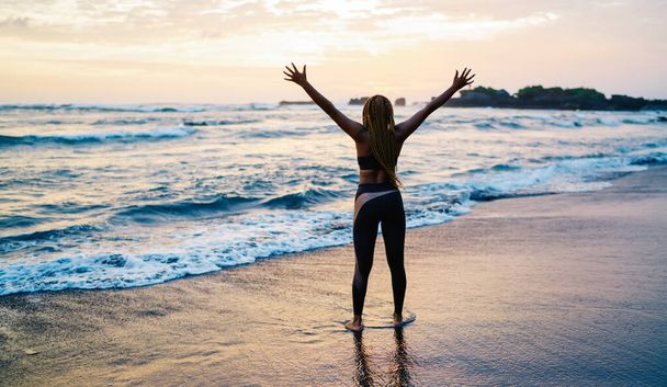 Back view of satisfied female runner in sportive tracksuit raising hands during workout break at seashore coastline, carefree woman with casual figure enjoying slimming goals and own triumph - Foto, Imagen