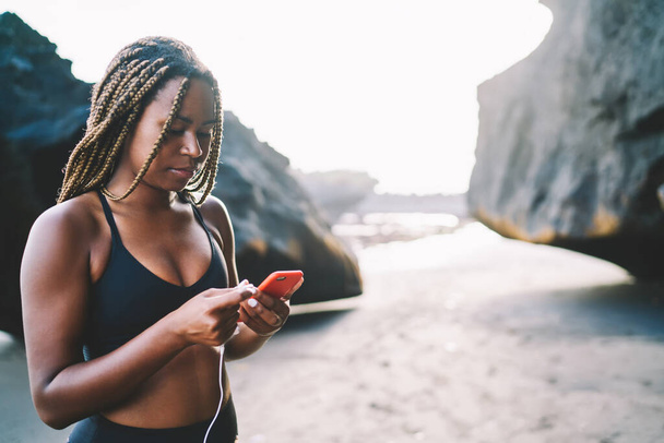 African American female in tracksuit checking received email spending morning for workout training at coastline beach, dark skinned fit girl in sportive clothing reading web publication on cellphone - Фото, зображення
