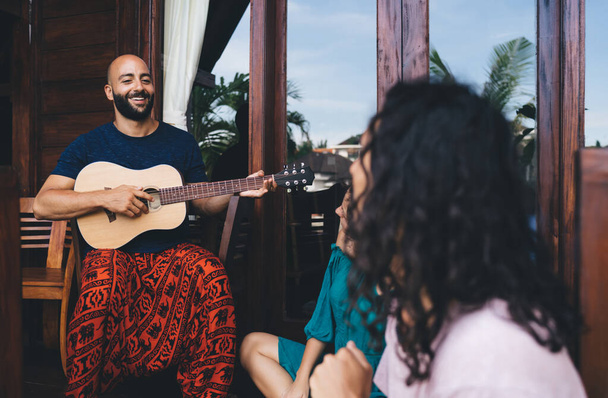 Group of happy multiethnic friends in casual clothes sitting on terrace and enjoying time together while playing guitar in sunny day - Foto, afbeelding