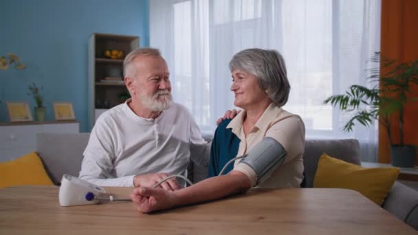 idős boldog pár nézi a kamerát vérnyomásmérés közben, férj használ vérnyomás monitor felesége magas vérnyomás otthon - Felvétel, videó
