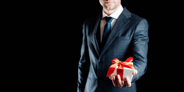 Gift man. Happy young businessman holding surprise gift box present with red ribbon isolated on black background. Black Friday, Valentine Day congratulations banner - Фото, изображение
